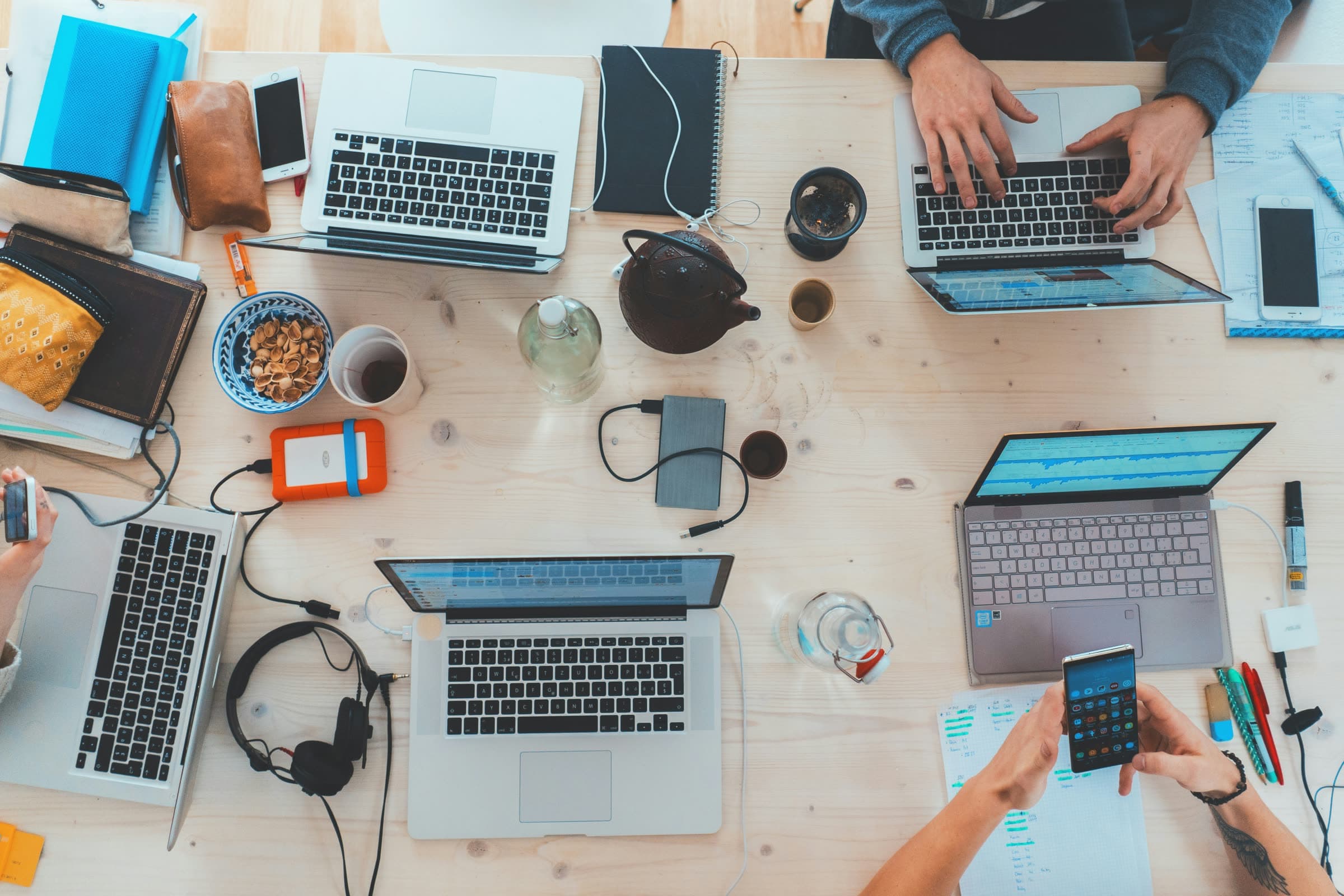 A shared studio desk shot from above — four laptops, notebooks, headphones and phones mid-session